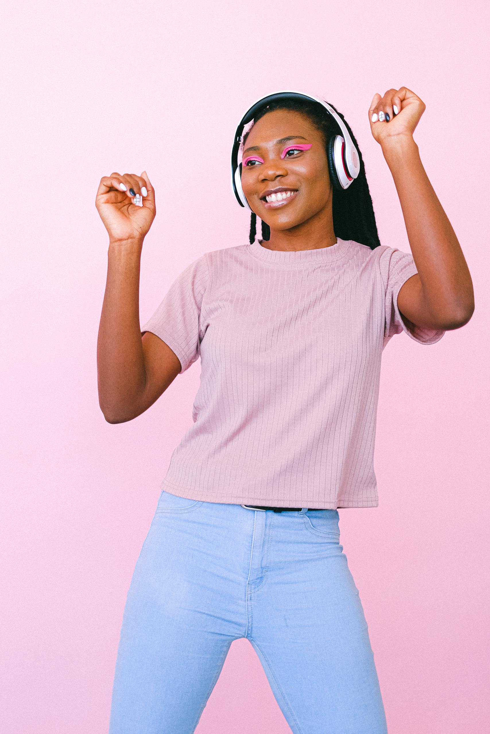 A young woman dances joyfully to music with headphones on a pastel pink background.